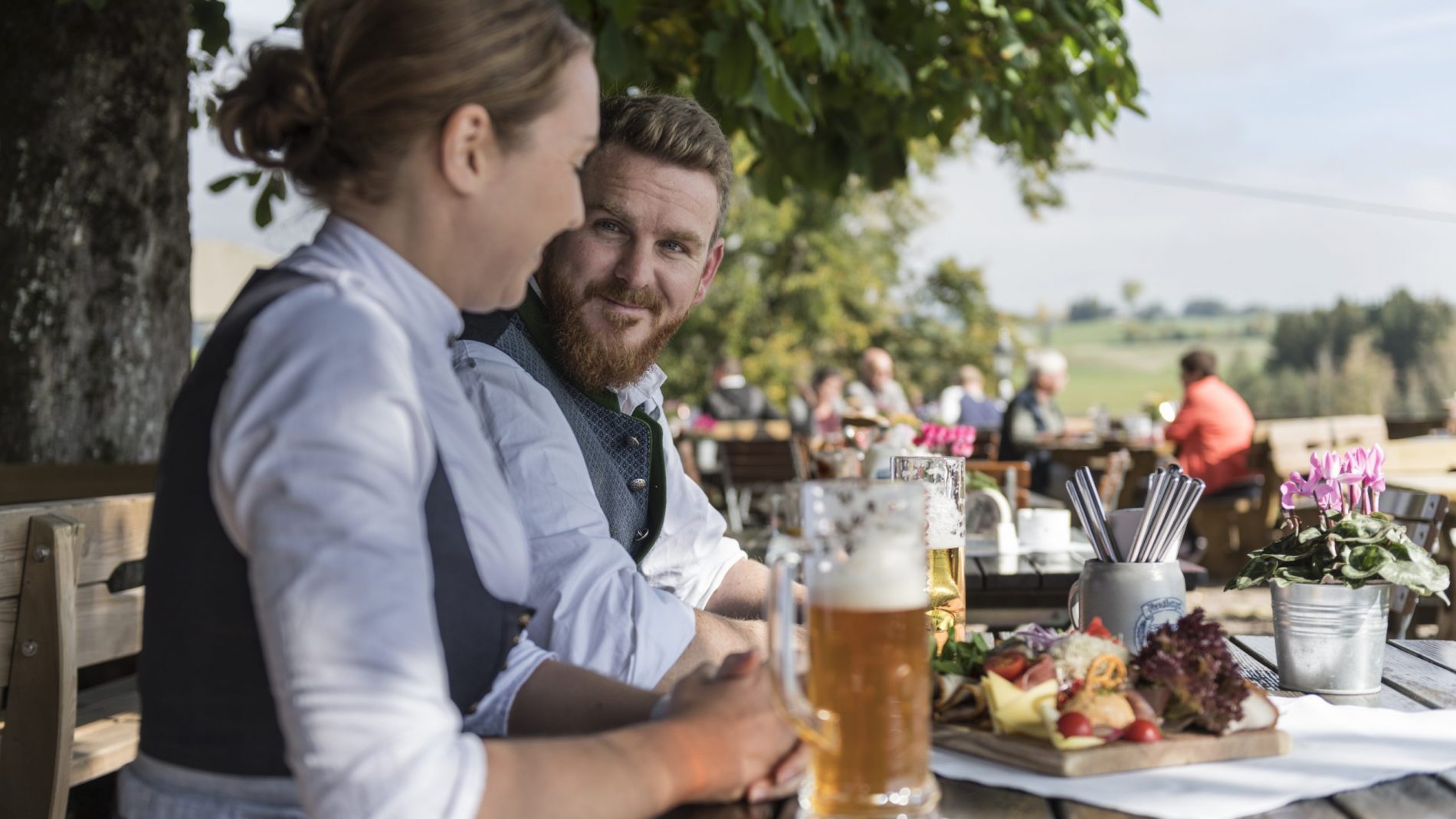Paar in traditioneller Kleidung sitzt im Biergarten, vor ihnen Bier und Käseplatte; Hintergrund weitere Gäste.