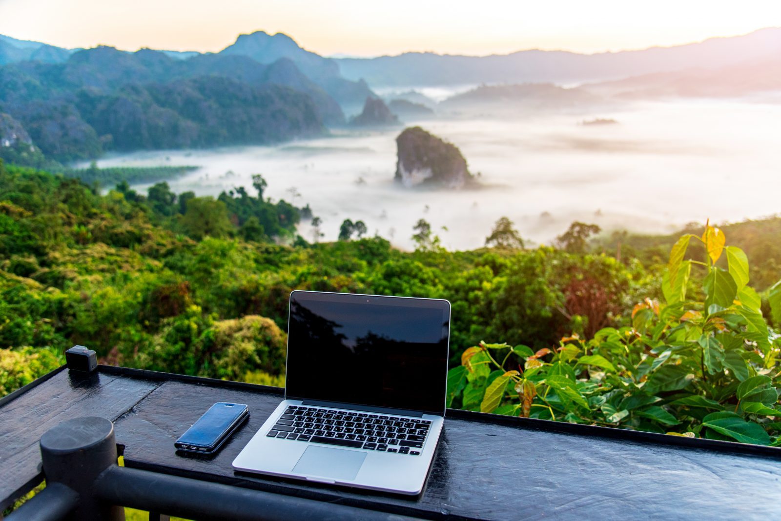 Ein Laptop steht auf einem breiten Balkon-Geländer, dahinter ein Weitblick auf Natur und Berge.