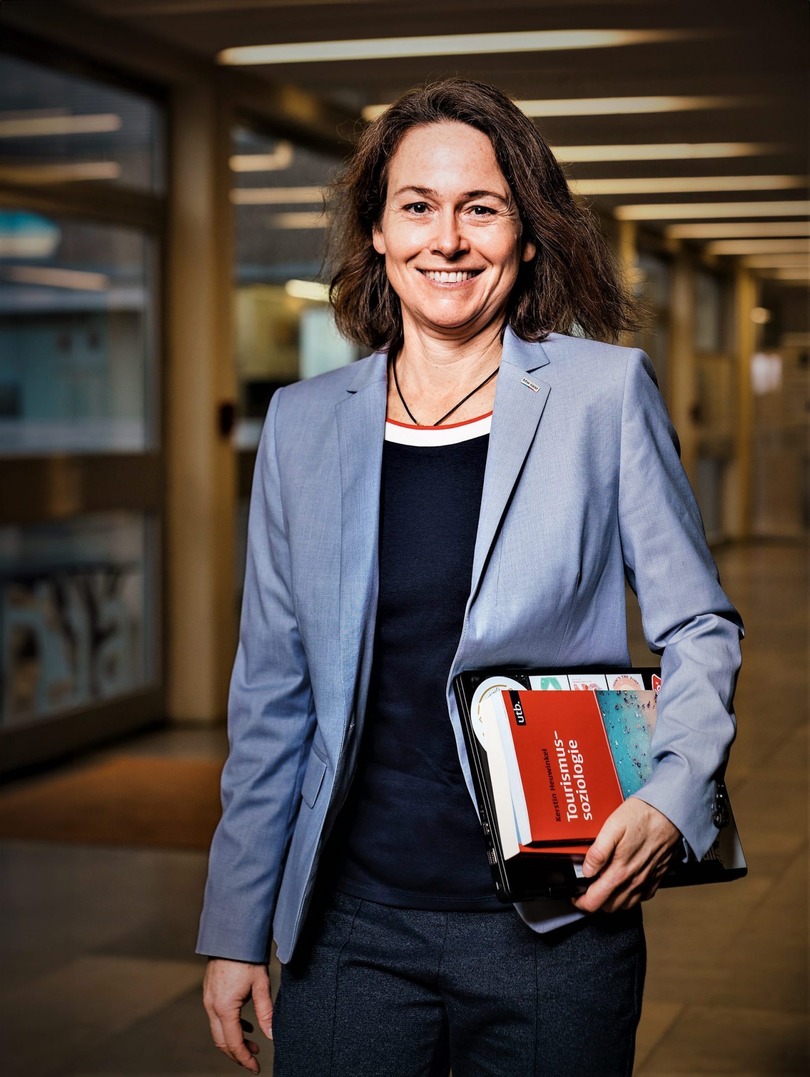 Kerstin Heuwinkel Kerstin Heuwinkel steht im Büroflur, trägt blaues Sakko und dunkelblaues Top, hält einen Ordner mit rotem Buch