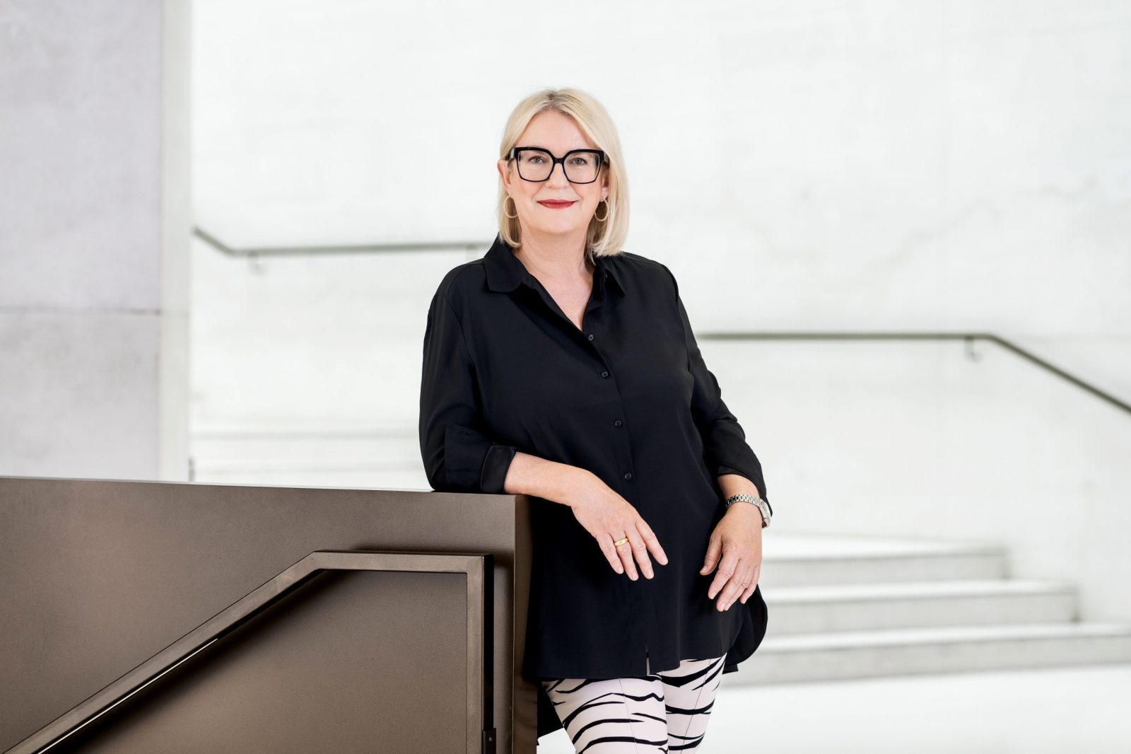 Petra_Hedorfer-9165 -Credit_DZT_Farideh Diehl Portrait einer blonden Frau mit schwarzen Brillengläsern, Bluse und gestreifter Hose lehnt an Geländer im hellem Treppenhaus.