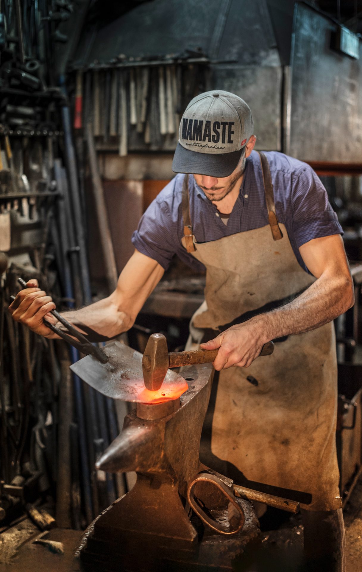 Ein Schmied bearbeitet glühendes Metall auf einem Amboss in einer traditionellen Schmiede, umgeben von Werkzeugen und Metallobjekten.