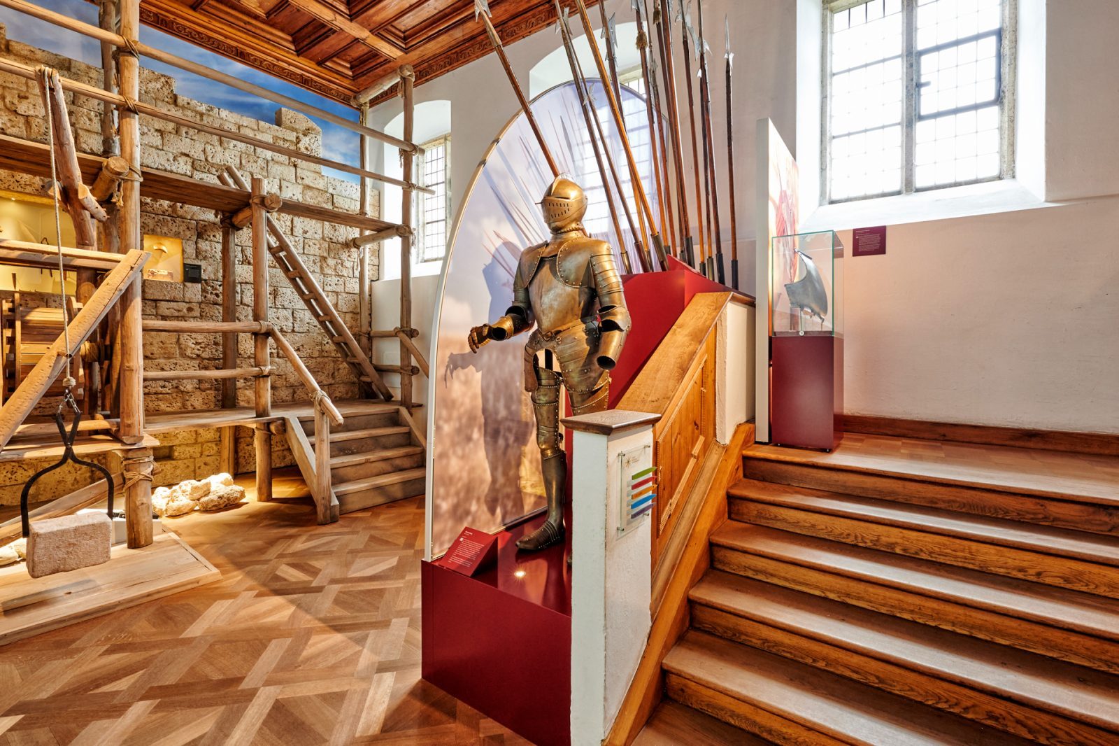 Innenansicht eines Museums mit einer Rüstung und historischen Waffen, die an einer Wand aus alten Steinen ausgestellt sind.