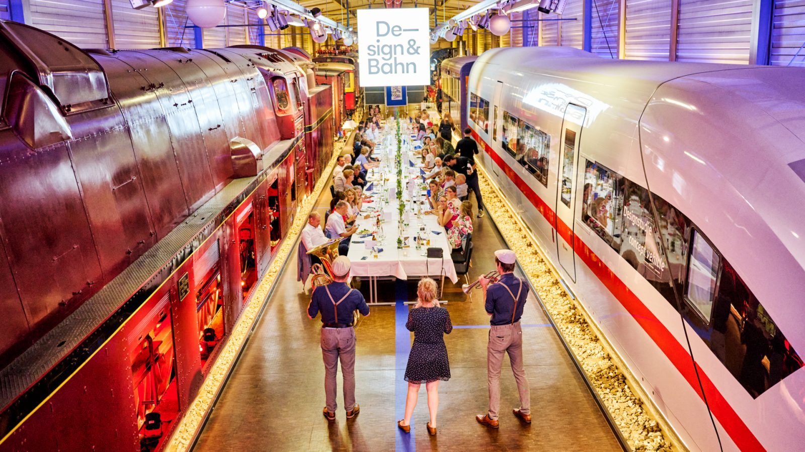 Festliches Abendessen im DB Museum Nürnberg mit Gästen an einem langen Tisch, umgeben von historischen und modernen Zügen. Musiker spielen in der Nähe.