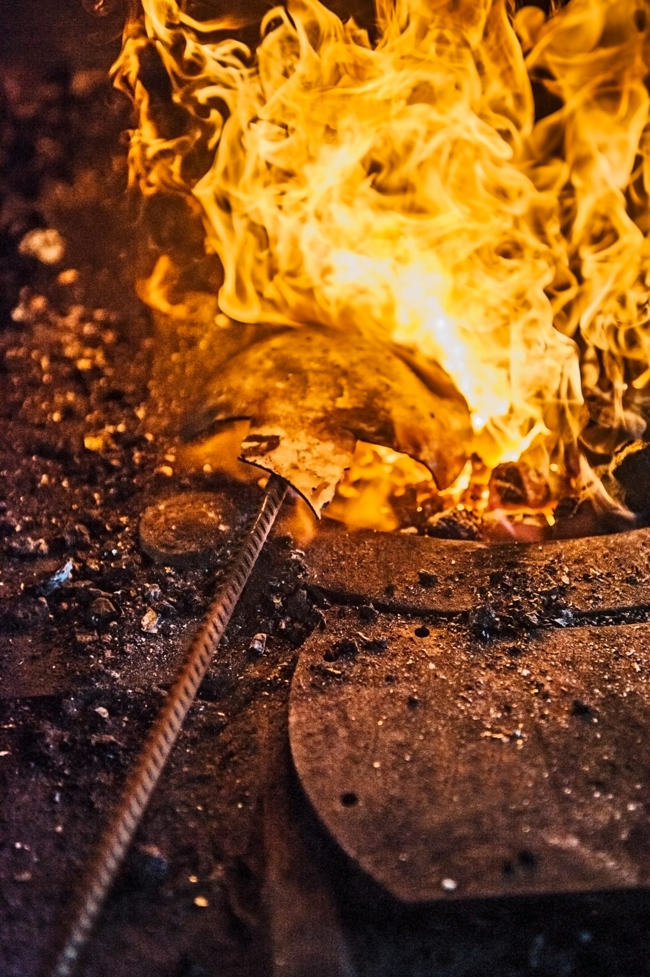 Eine Nahaufnahme eines Schmiedefeuers mit Flammen und glühendem Metall, umgeben von glühenden Kohlen und Werkzeugen.
