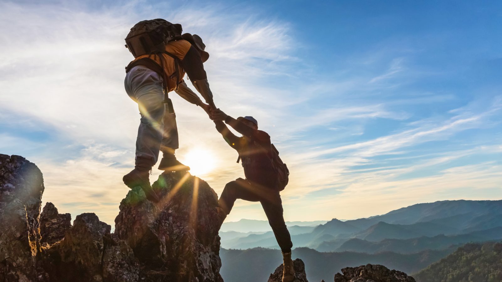 Zwei Bergsteiger helfen sich beim Klettern auf einem Felsen, mit einer malerischen Berglandschaft im Hintergrund und der Sonne am Horizont.