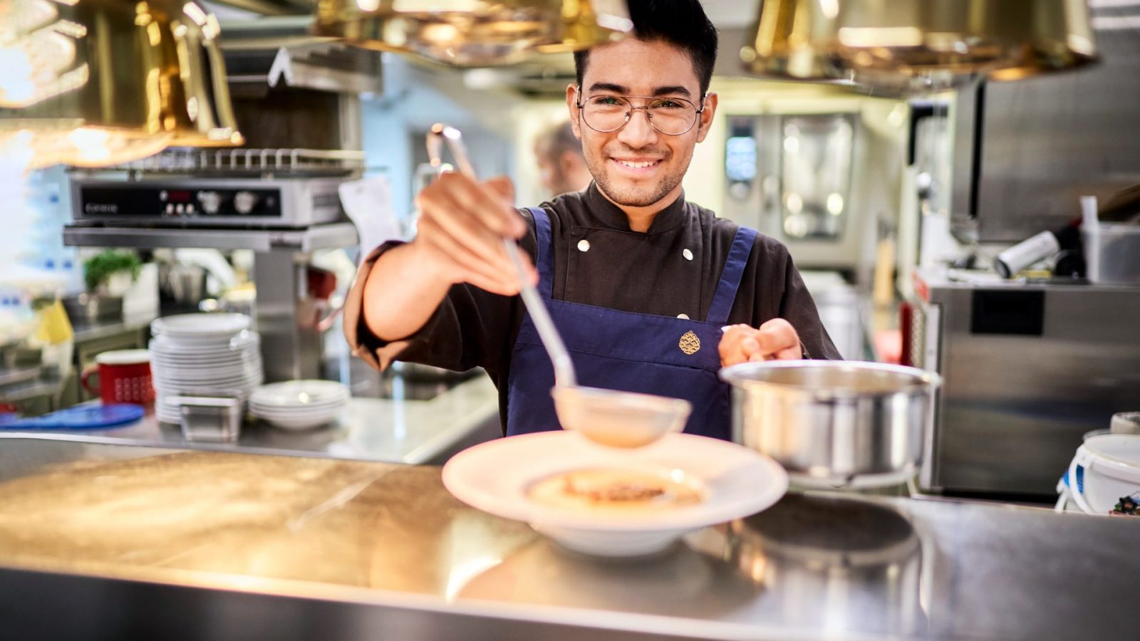 Ein Auszubildender in einem Restaurant löffelt eine Suppe in eine weiße Schüssel und lächelt dabei. Im Hintergrund sind Küchengeräte und Geschirr zu sehen.