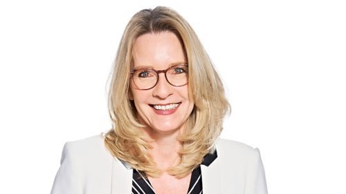 Eine lächelnde Frau mit blonden, langen Haaren und Brille, die in einer formellen Bluse und Jacke gekleidet ist.