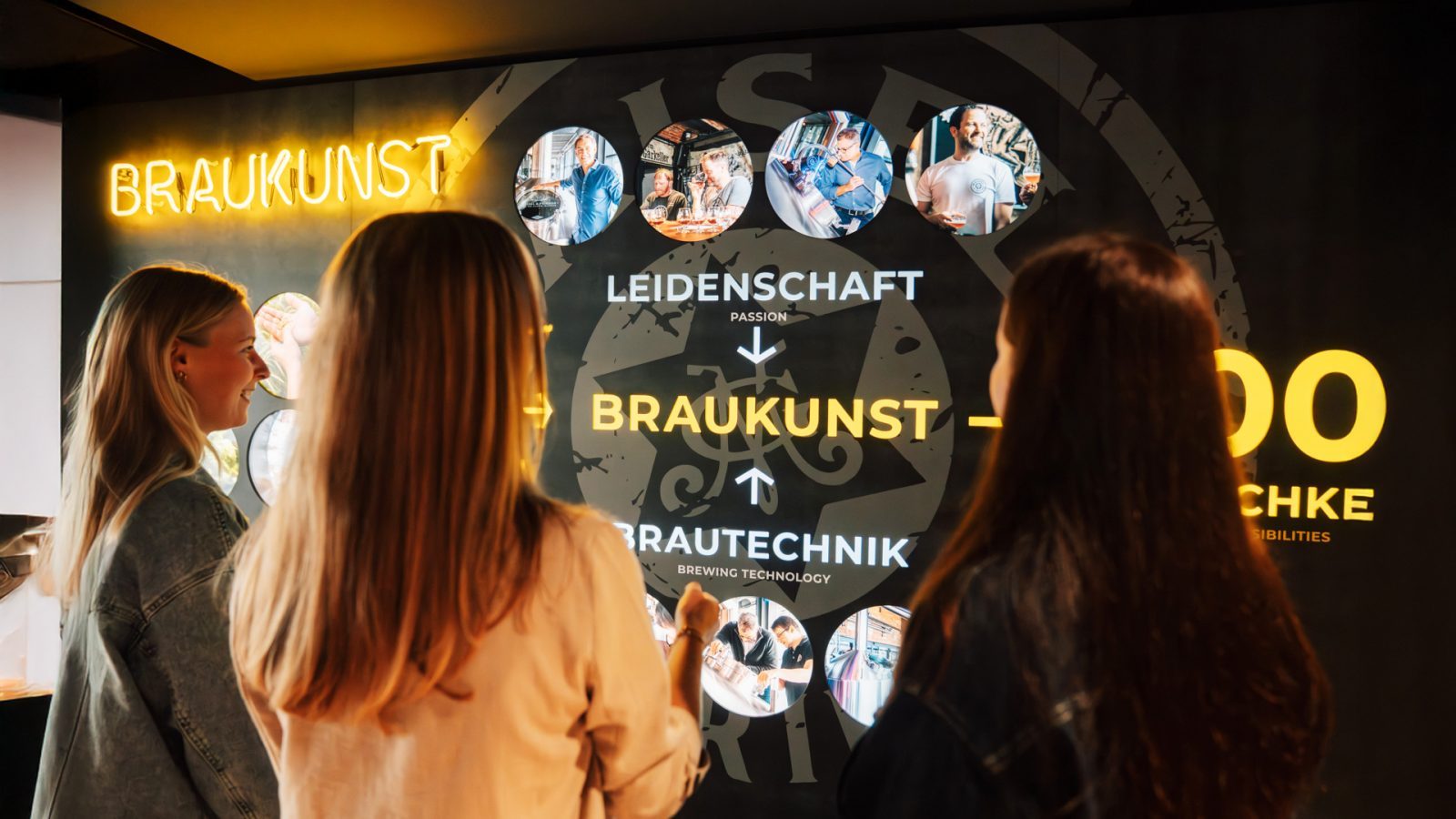 Zwei Frauen stehen vor einem beleuchteten Wanddisplay in der Braukunstwelt, das Informationen zur Brautechnologie und Leidenschaft für das Bierbrauen zeigt.