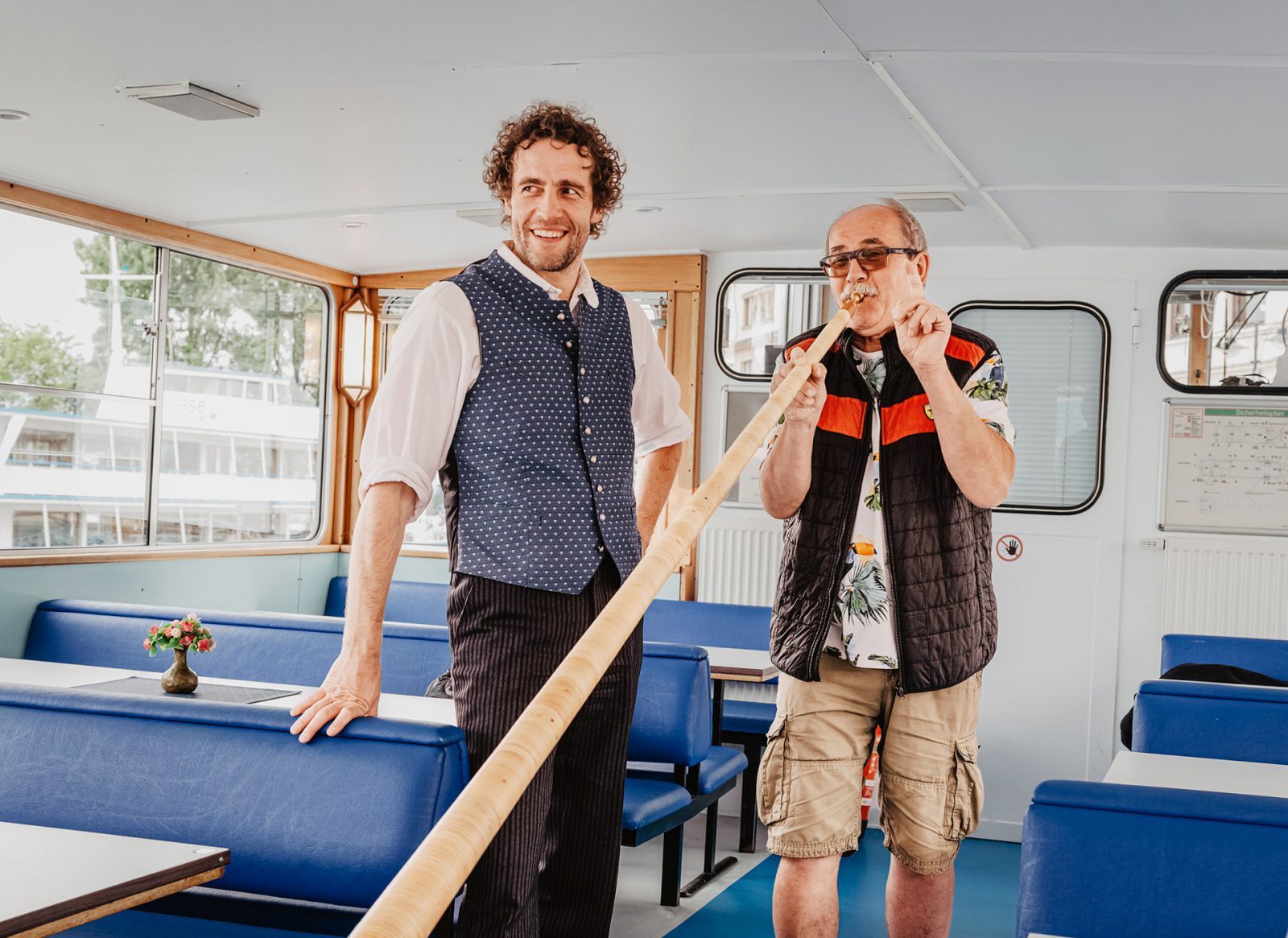 Zwei Männer stehen in einem Boot. Einer spielt auf einem Alphorn, der andere lächelt. Das Innere des Bootes ist hell und freundlich gestaltet.