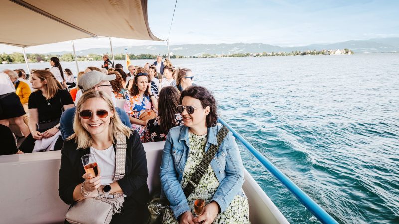 Zwei Frauen auf einem Boot auf dem Bodensee, umgeben von anderen Passagieren. Eine hält ein Glas Sekt. Hintergrund zeigt den See und die Uferlandschaft.