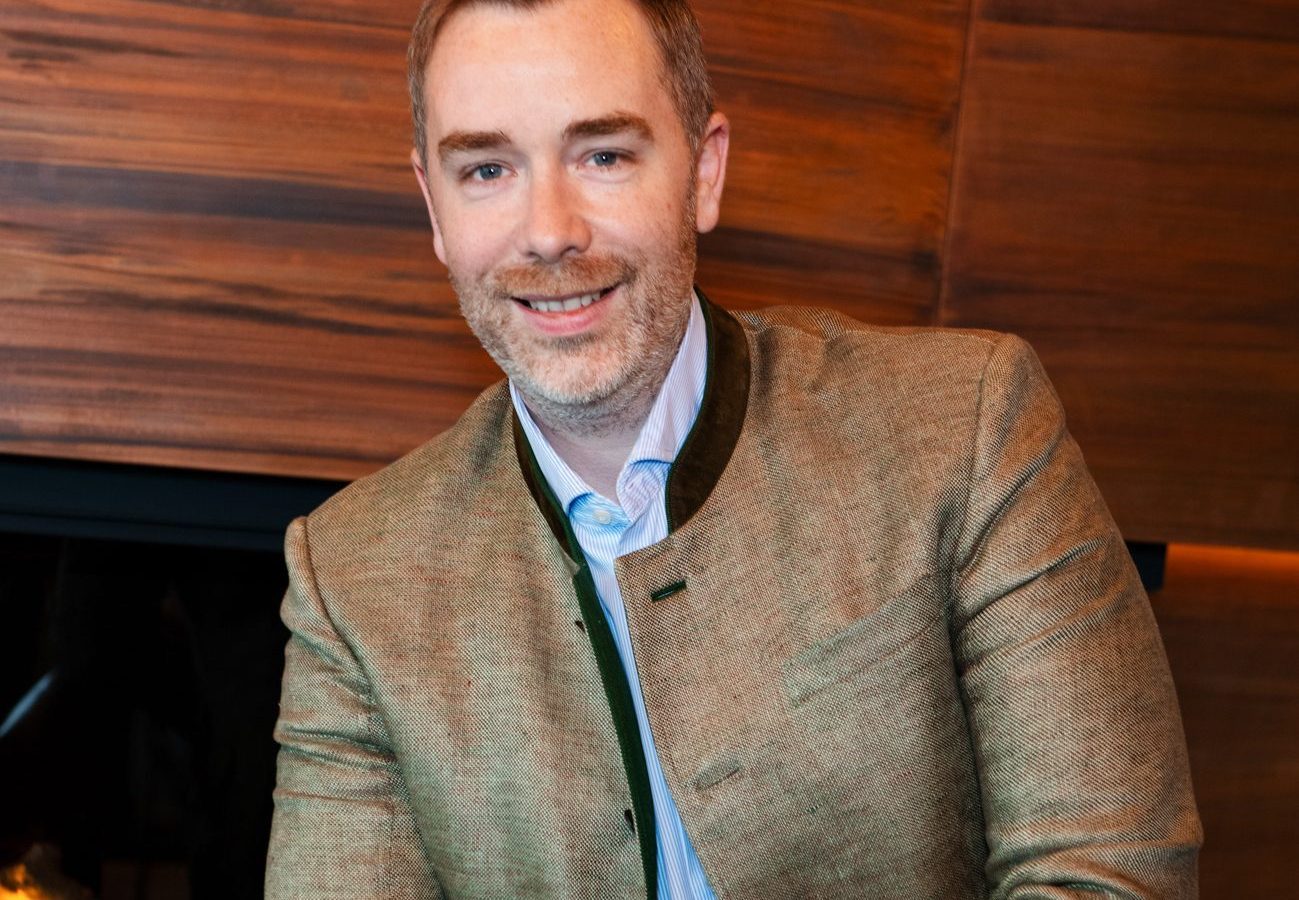 Porträt eines Mannes in einem stilvollen Hotelambiente, mit einem Kamin im Hintergrund und einer warmen Holzfassade. Der Mann trägt ein blaues Hemd und eine hellbraune Jacke.