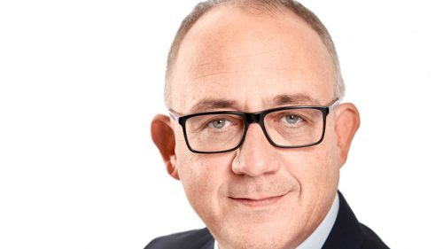 Porträt von einem Mann mit Brille und kurzen Haaren. Er trägt ein Jackett, Hemd und Krawatte, der Hintergrund ist weiß.