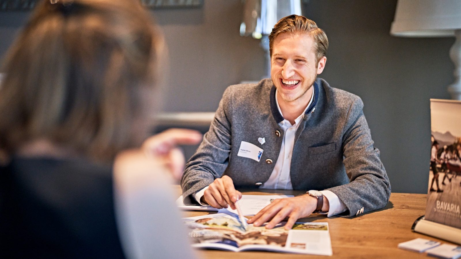 Ein Mann in traditioneller Tracht lächelt am Tisch und zeigt auf ein Prospekt, während eine Frau ihm gegenübersitzt.