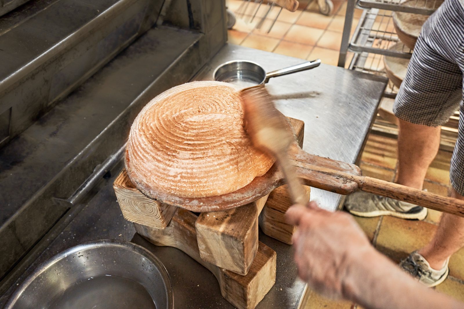 Eine Person formt einen runden Brotlaib auf einem Holzbrett in einer Backstube. In der Umgebung sind Werkzeuge und Utensilien sichtbar.
