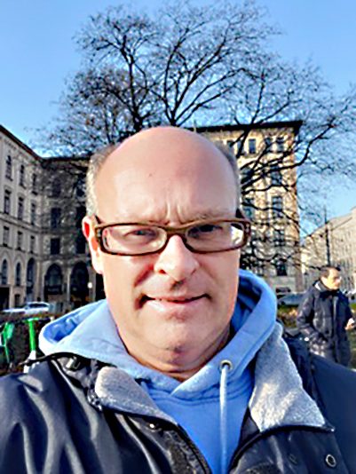 Ein Mann mit Brille und Kapuzenpullover steht in einer städtischen Umgebung, umgeben von Bäumen und Gebäuden.