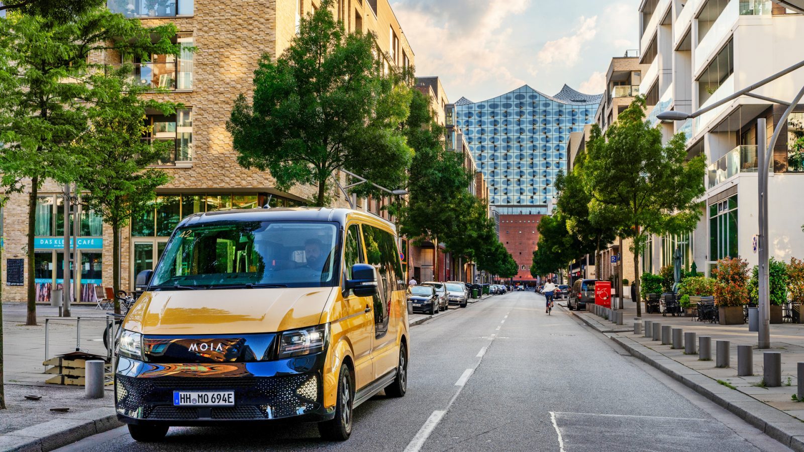 Ein gelber MOIA-Ridesharing-Bus steht auf einer Straße in Hamburg, umgeben von modernen Gebäuden und Grünanlagen. Im Hintergrund ist die Elbphilharmonie zu sehen.