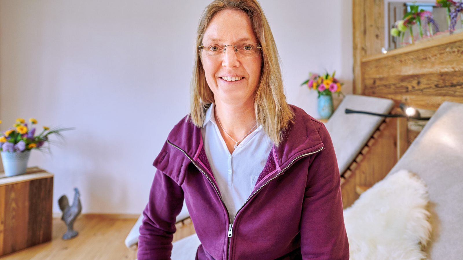 Eine Physiotherapeutin sitzt in ihrer Praxis und lächelt freundlich. Im Hintergrund sind eine weiche Liege, Holzverkleidungen und Pflanzen dekoriert.