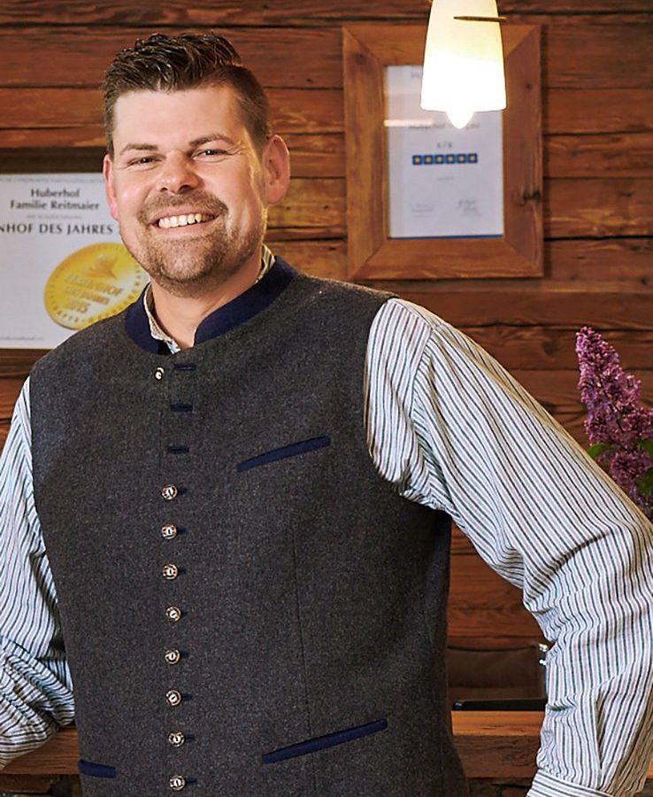 Ein Mann in traditioneller Tracht steht lächelnd vor einer Holztheke, umgeben von rustikalem Interieur.