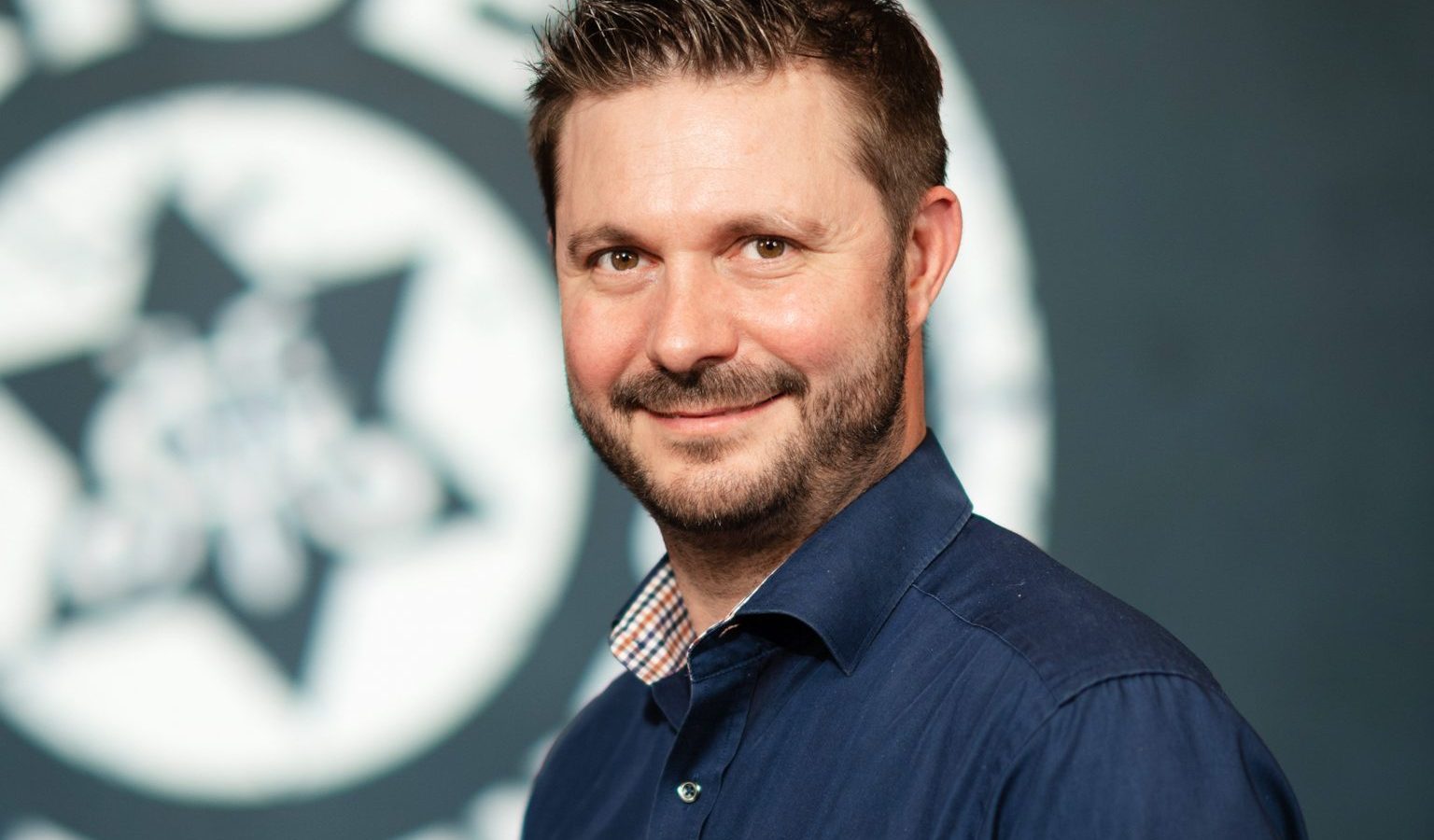 Porträt von Michael Feuerstein, Geschäftsführer der Bayreuther Bier-Erlebniswelt, vor einem Hintergrund mit dem Logo der Brauerei.