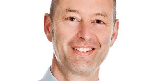 Porträt eines Mannes mit Glatze, der ein Hemd und eine Krawatte trägt, lächelnd vor weißem Hintergrund.