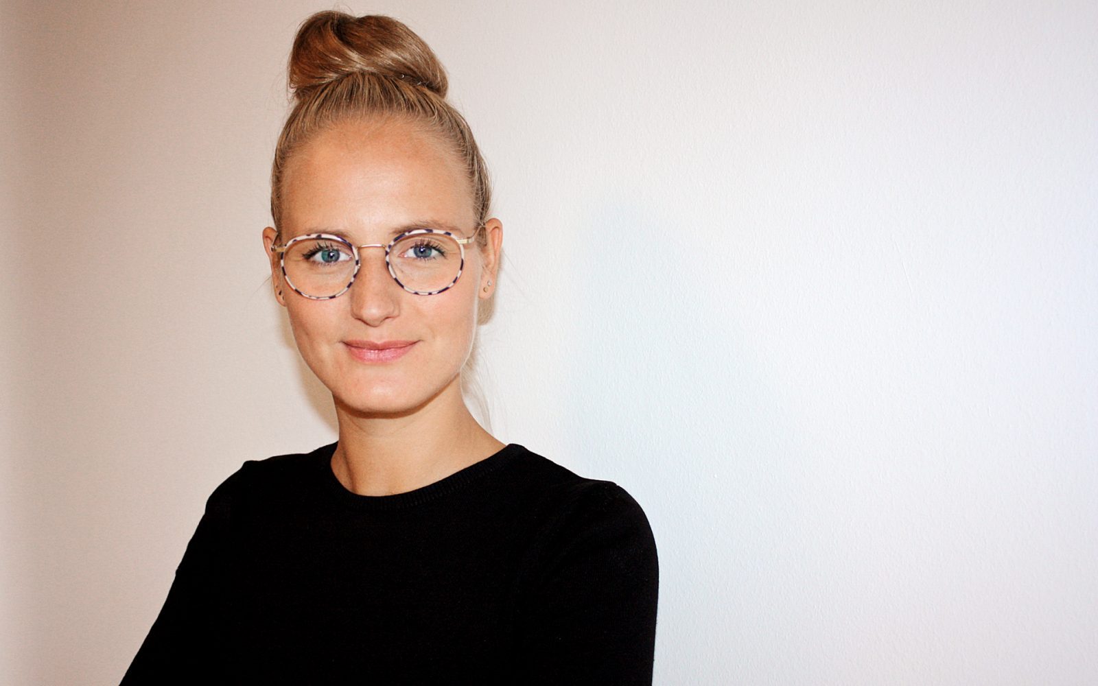 Portrait von Birthe Menke, einer Frau mit blonden Haaren in einem hohen Dutt, trägt eine schwarze Bluse und eine modische Brille, vor einem hellen Hintergrund.
