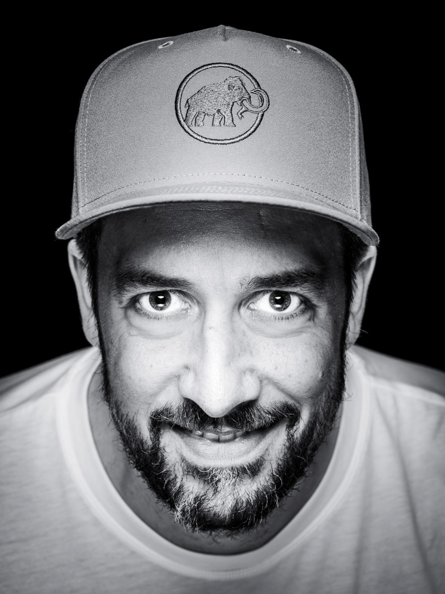 Nahaufnahme eines Mannes mit Bart und Baseballcap, der in die Kamera lächelt, vor schwarzem Hintergrund.