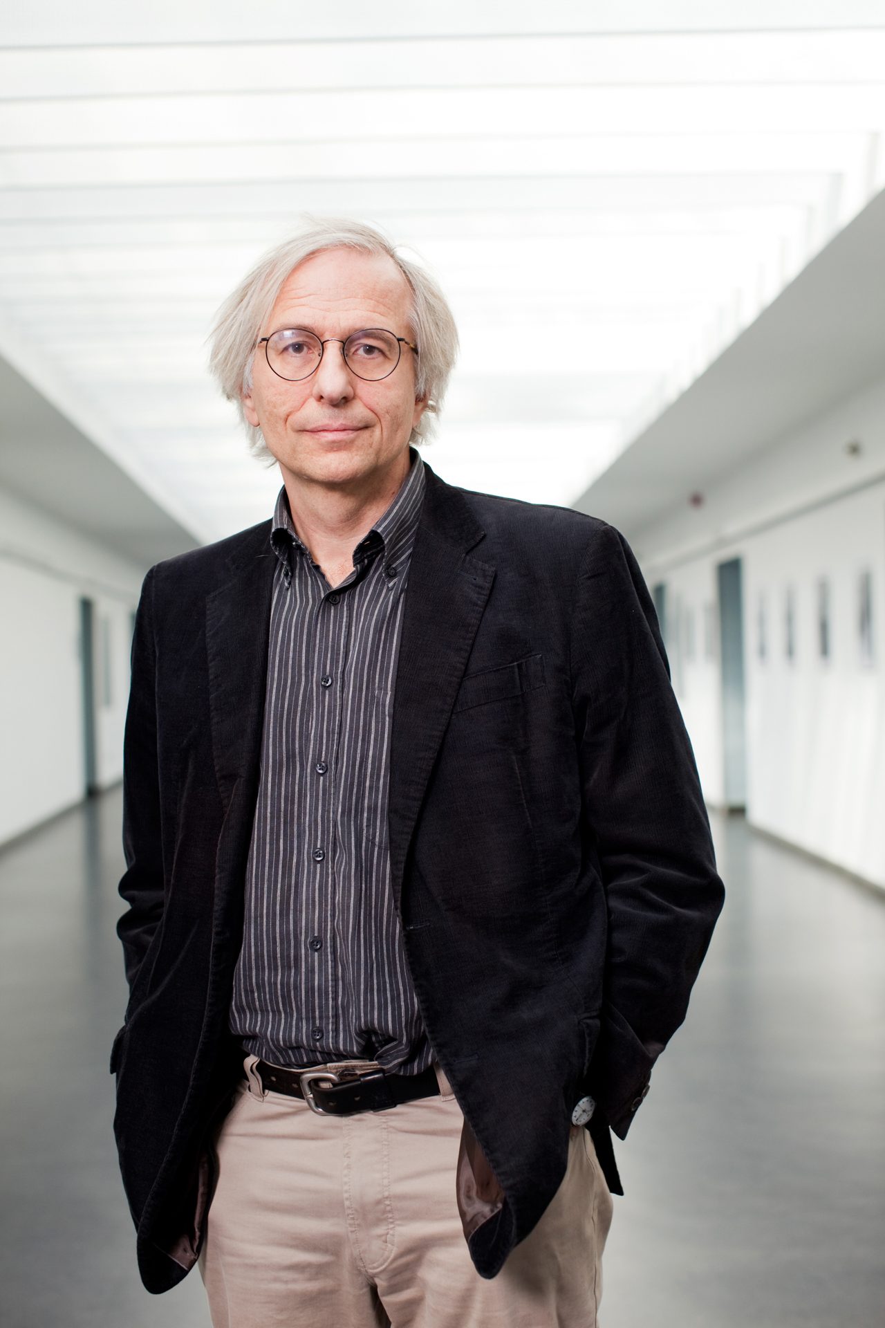 Ein Mann mit grauen Haaren und runder Brille steht in einem modernen, lichtdurchfluteten Flur eines Gebäudes und blickt zur Kamera.