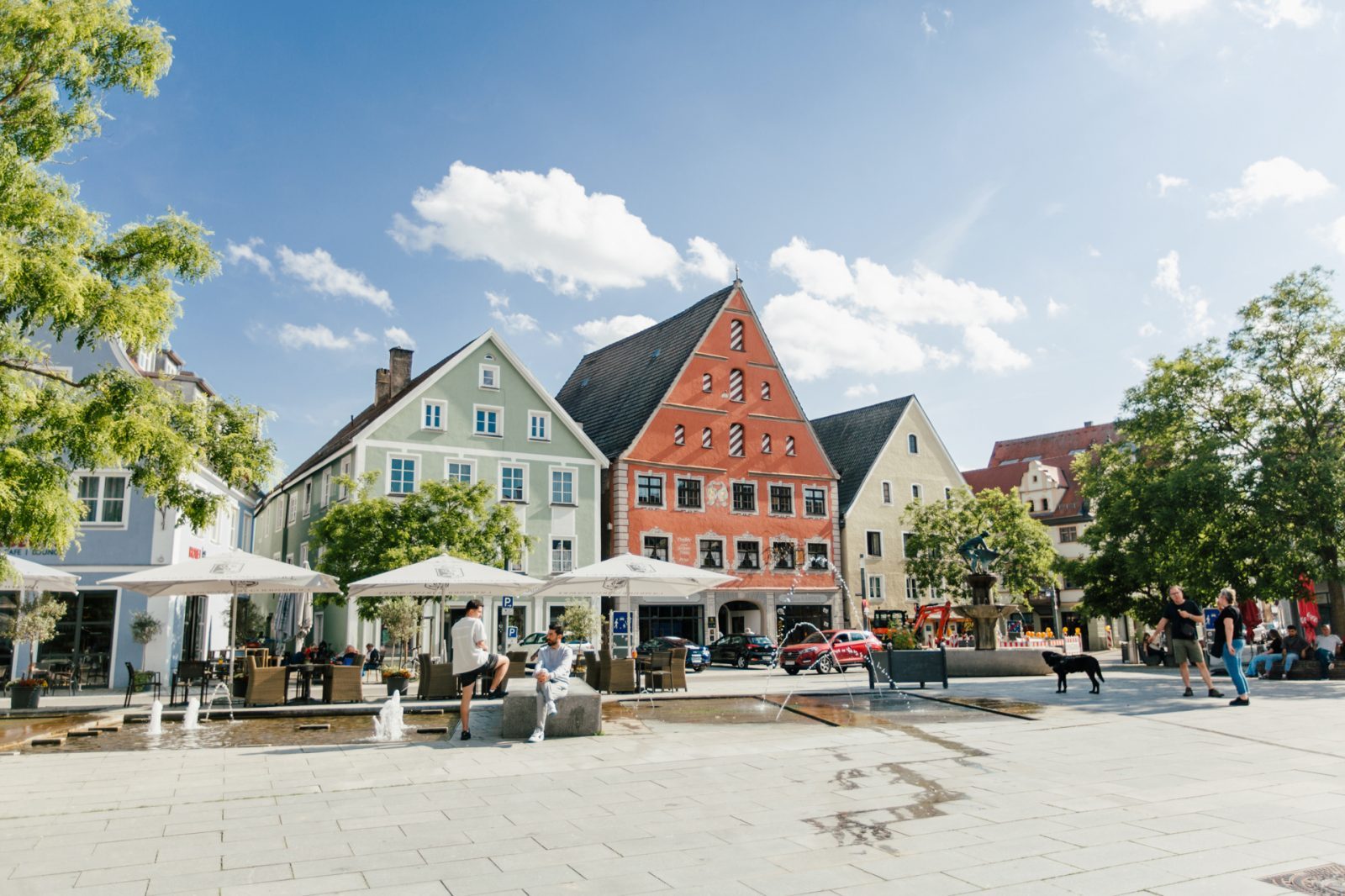 Der Schrannenplatz mit bunten Häusern und Menschen, die sich unter Sonnenschirmen entspannen, und Wasserspielen in einer angenehmen Atmosphäre.