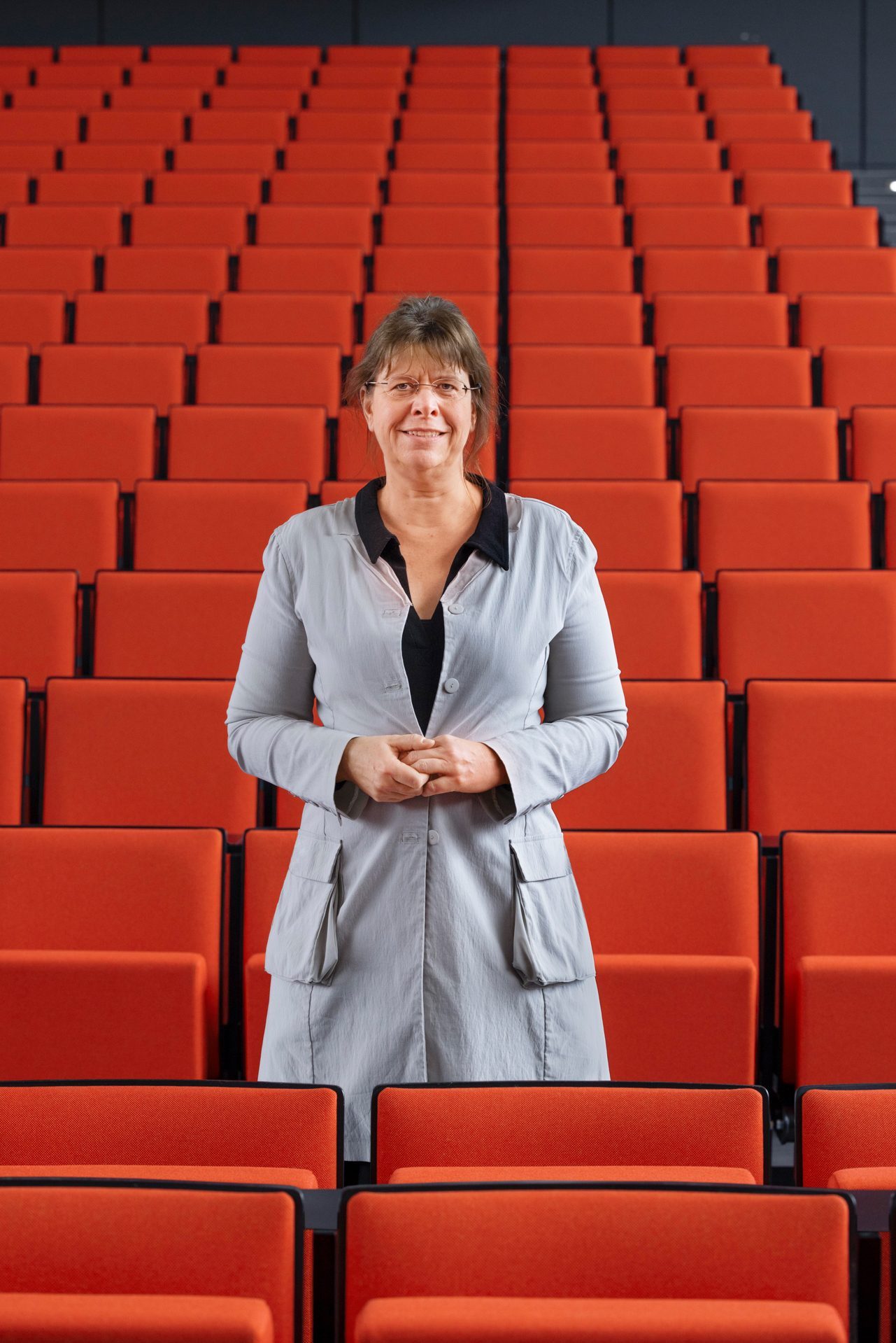 Eine Frau in hellgrauer Jacke steht in einem leeren Theaterraum mit rot gepolsterten Sitzen.
