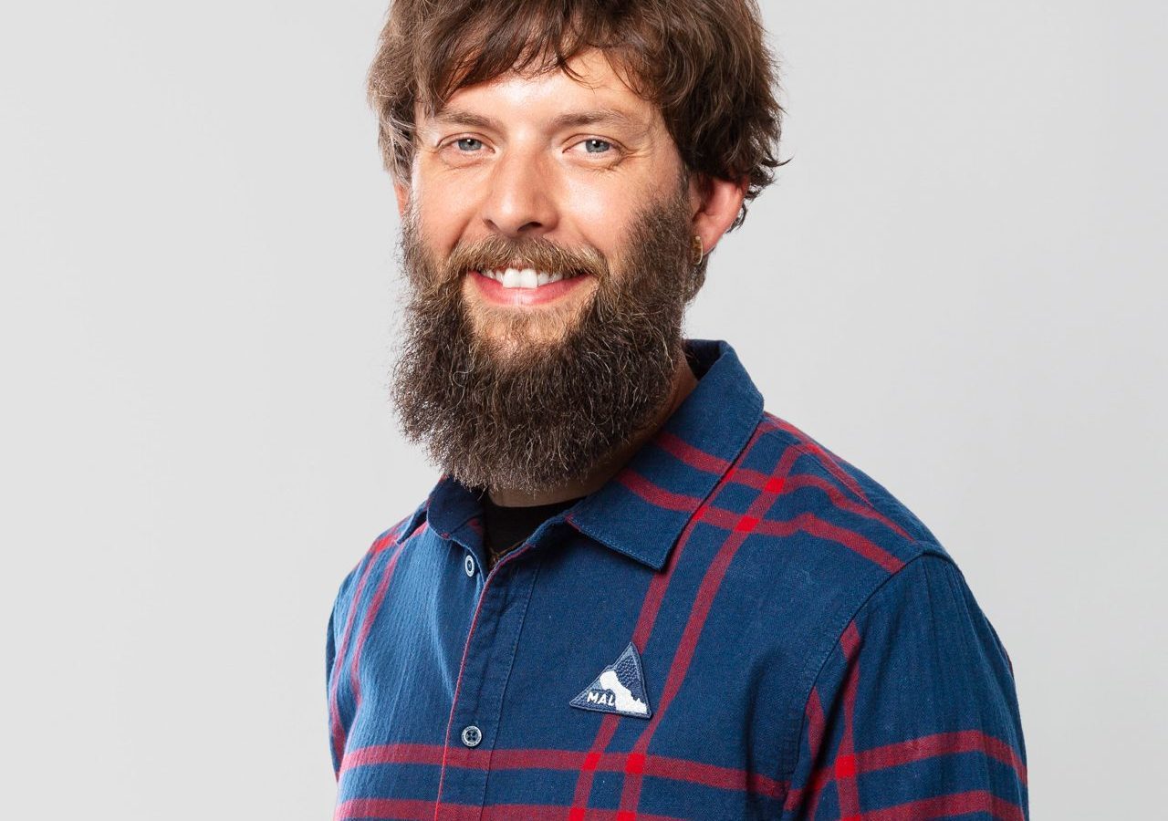 Freundlicher Mann mit Bart und kariertem Hemd, steht vor neutralem Hintergrund und lächelt.