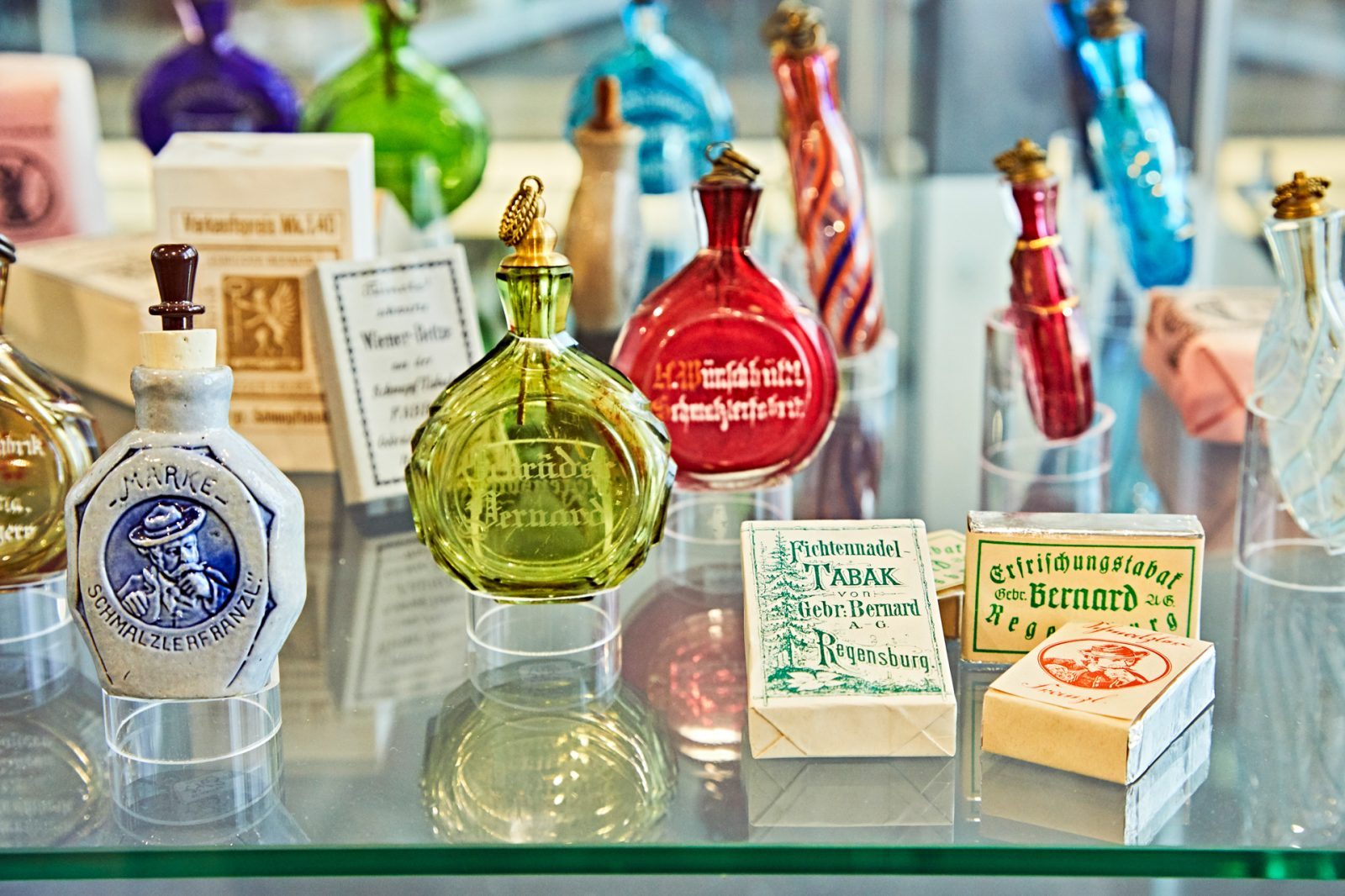 Eine Vitrine zeigt verschiedene alte Glasflaschen und Verpackungen aus der bayerischen Glasbläserei, darunter farbige Flaschen und Zigarettenschachteln.