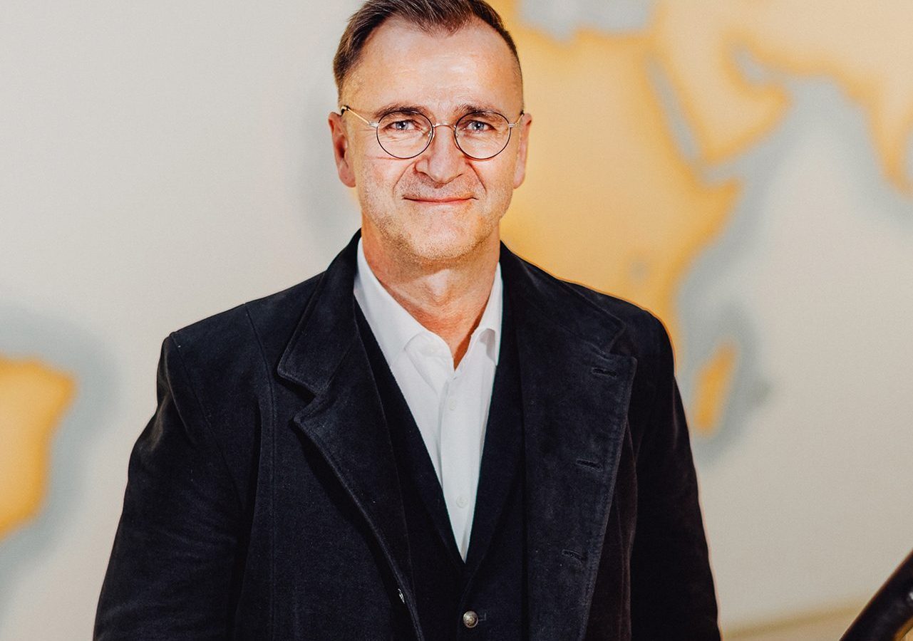 Portait eines Mannes in schwarzem Anzug, stehend vor einer Weltkarte in sanften Farben.