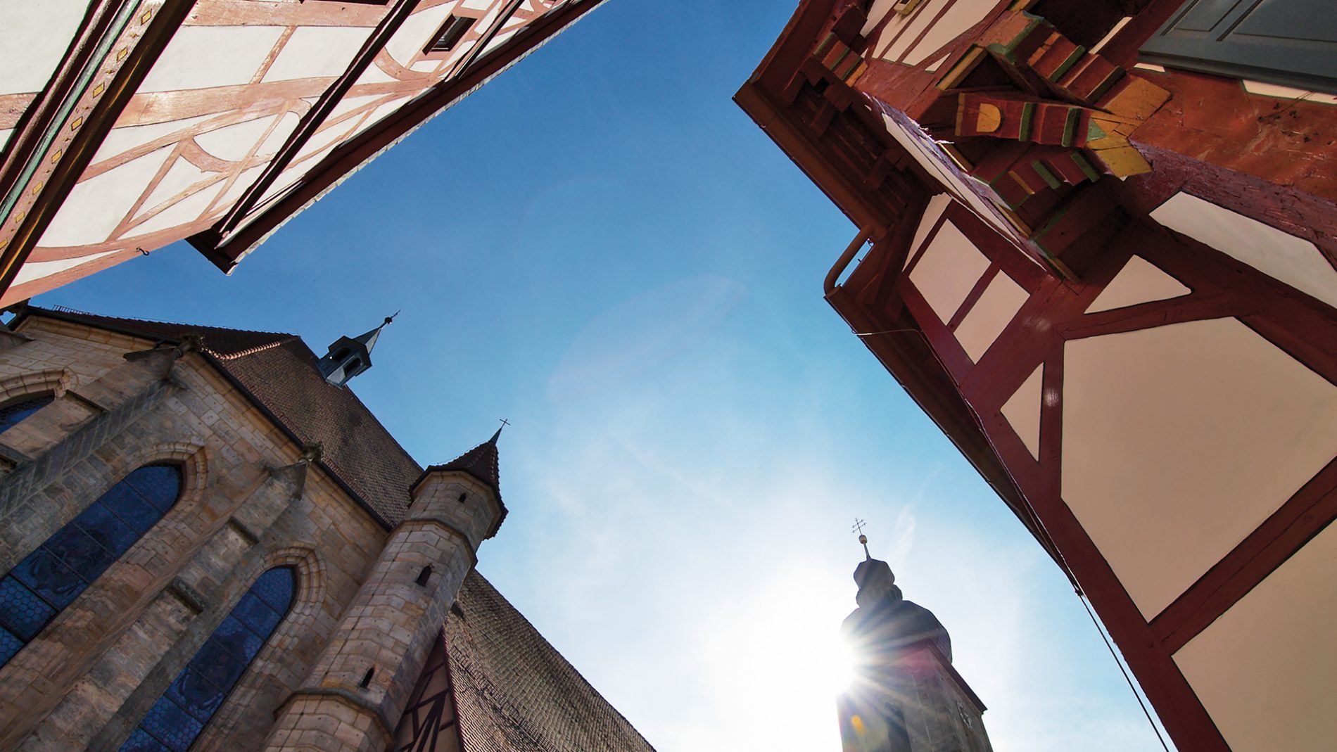 Blick auf Fachwerkhäuser und eine Kirche unter strahlend blauem Himmel. Die Sonnenstrahlen scheinen zwischen den Gebäuden hindurch.