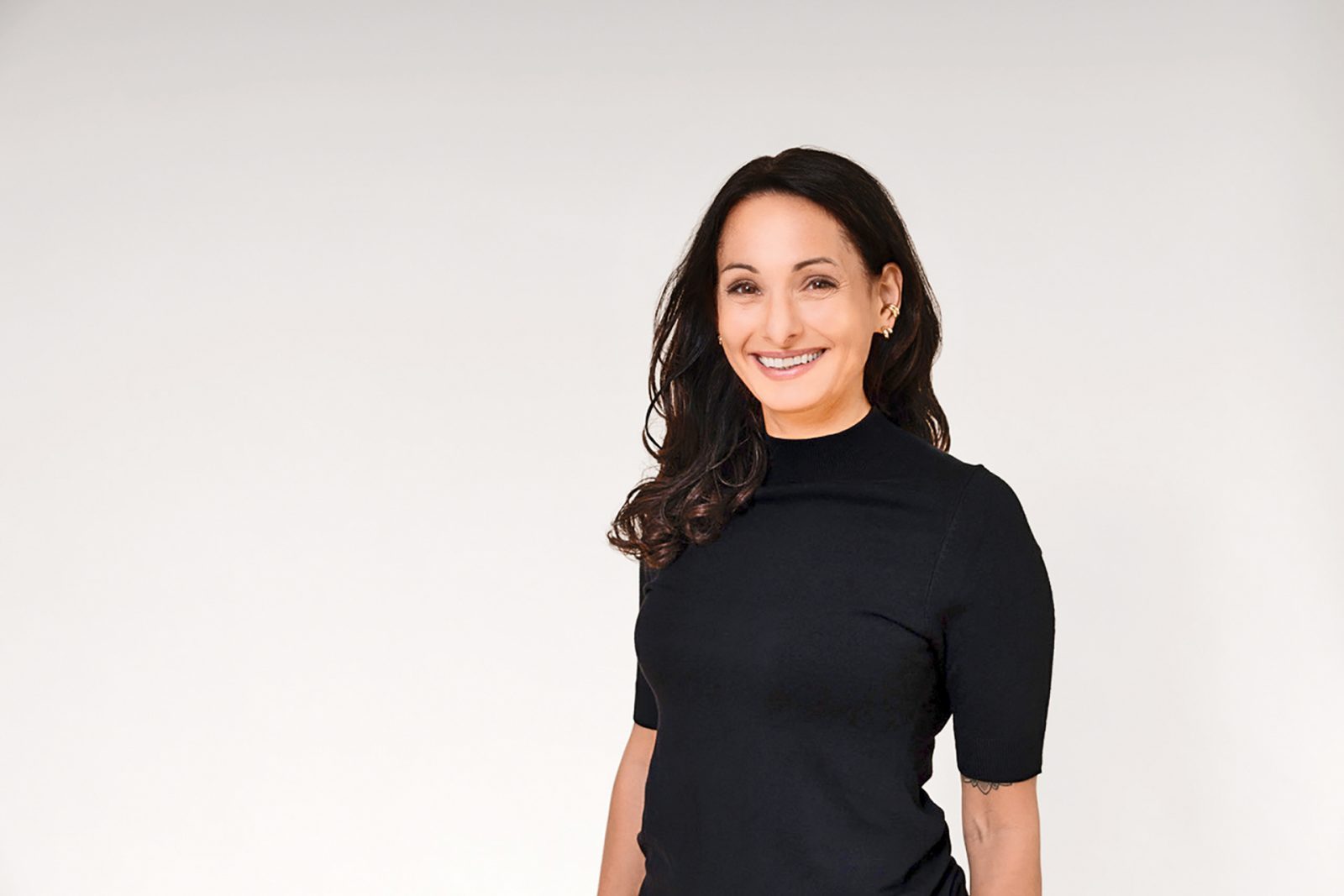 Porträt einer lächelnden Frau in schwarzem Oberteil vor einfarbigem Hintergrund.