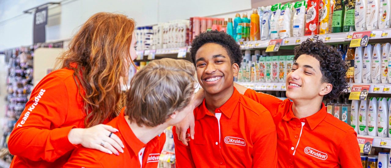 Vier Jugendliche in roten Kruidvat-T-Shirts lächeln und stehen vor einem Regal mit Pflegeprodukten.