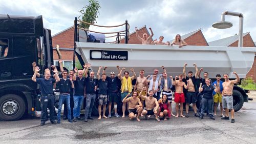 Gruppenszene mit 25 Personen vor einem Lkw, dessen Anhänger eine riesige Badewanne ist, alle jubeln und haben die Arme erhoben.
