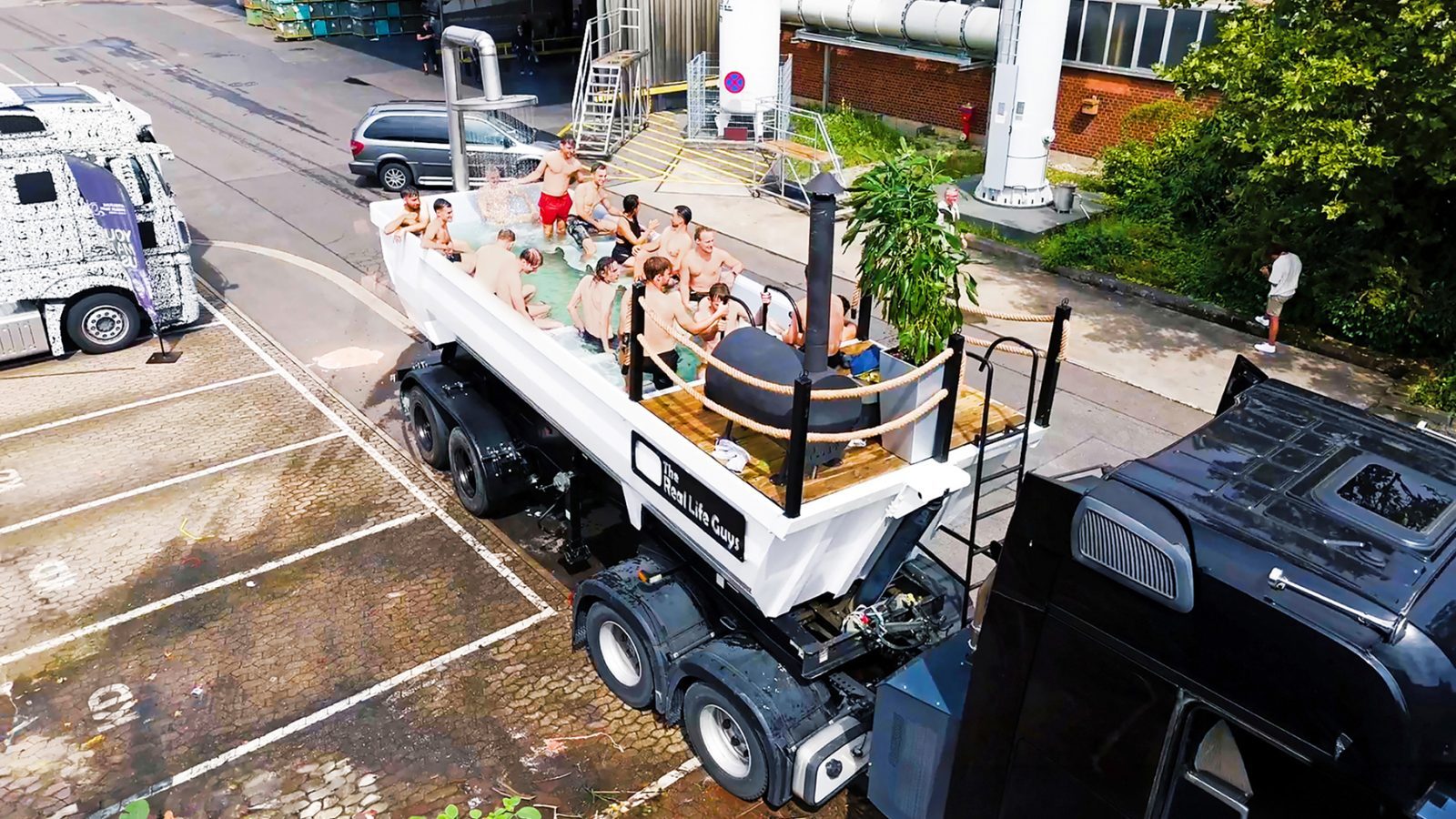 Eine Gruppe von Menschen sitzt in einem mit Wasser gefüllten Lkw-Anhänger in Form einer Badewanne auf einem Parkplatz.