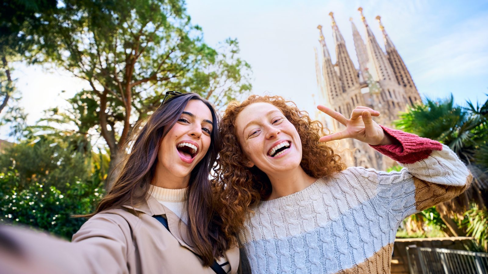 Zwei lachende Frauen machen ein Selfie vor der Sagrada Família mit Bäumen und Grünpflanzen im Hintergrund.