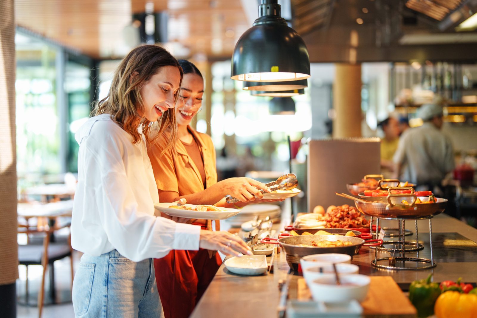 AdobeStock_1215219756 Zwei Frauen nehmen sich Essen an einem Buffet in einem modernen Restaurant, mit mehreren Gerichten und Getränken im Hintergrund.