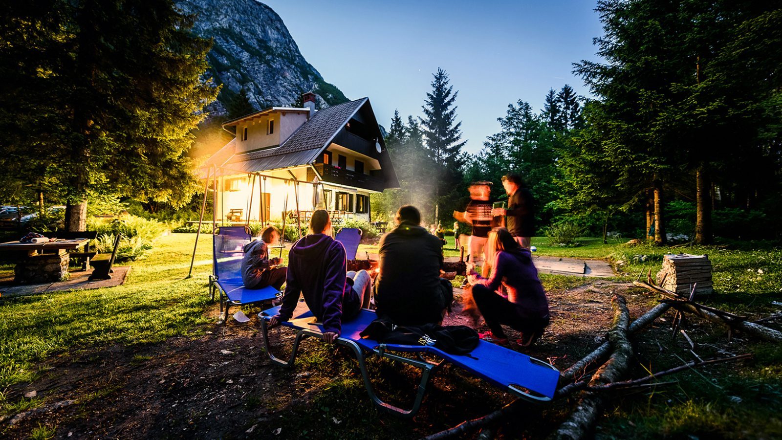 Freunde sitzen um ein Lagerfeuer vor einem Gebäude in der Natur, umgeben von Bäumen und einem klaren Abendhimmel.