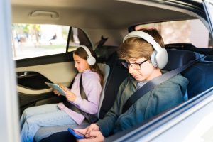 Zwei Kinder sitzen auf der Rückbank eines Autos, tragen Kopfhörer und nutzen Tablets. Hintergrund zeigt den Innenraum des Fahrzeugs.
