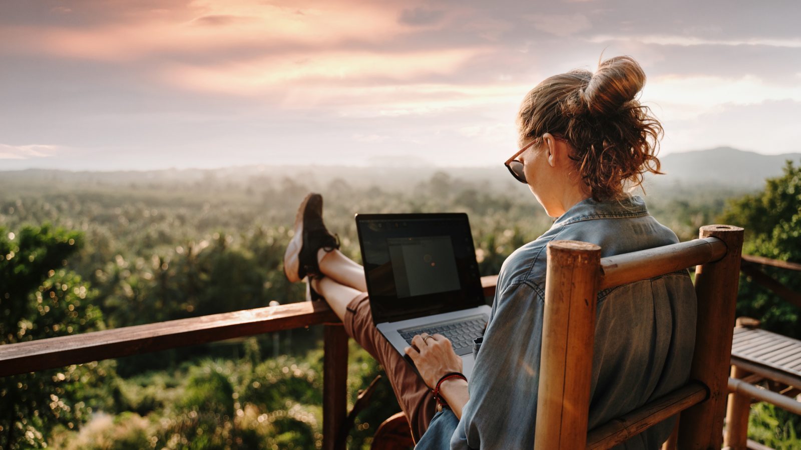 Frau sitzt auf einer Terrasse mit Laptop, blickt auf eine grüne Landschaft und einen bunten Sonnenuntergang.