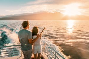 Paar steht auf einem Boot und betrachtet einen Sonnenuntergang über dem Wasser mit Bergen im Hintergrund.