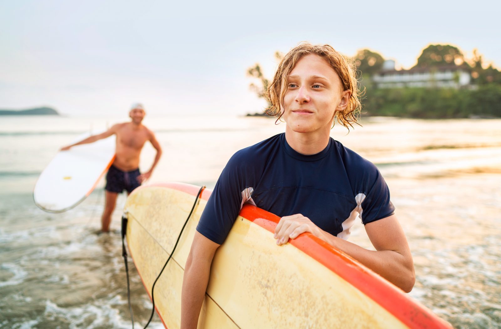 AdobeStock_480056562 Junger Surfer mit Surfbrett am Strand, im Hintergrund ein männlicher Surfer, sanfte Wellen und Sonnenuntergang.