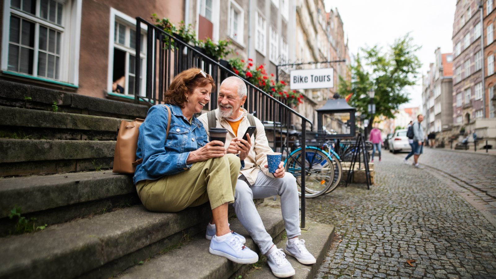 Ein lächelndes älteres Paar sitzt auf Stufen vor einem Gebäude in einer städtischen Straße, hält Kaffeebecher in der Hand und schaut auf ein Smartphone.