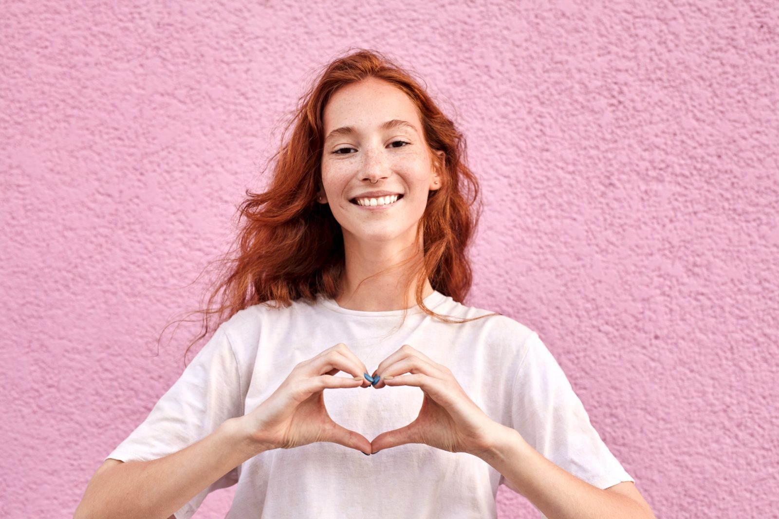 AdobeStock_518364404 Lächelnde Frau mit roten Haaren formt ein Herz mit ihren Händen vor einer rosa Wand.