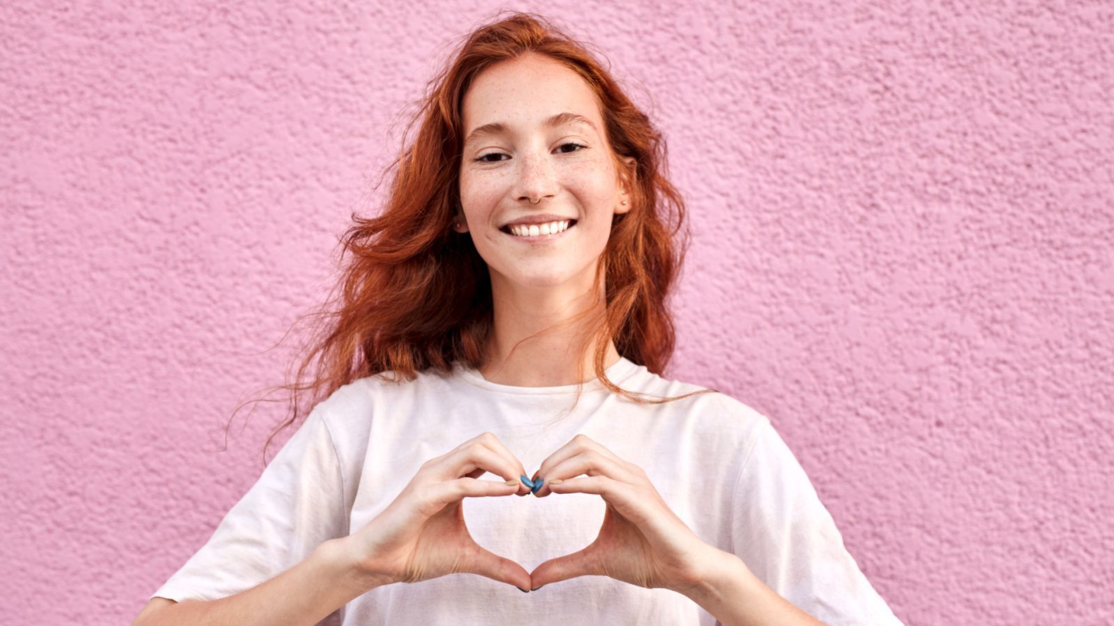 Lächelnde Frau mit roten Haaren formt ein Herz mit ihren Händen vor einer rosa Wand.