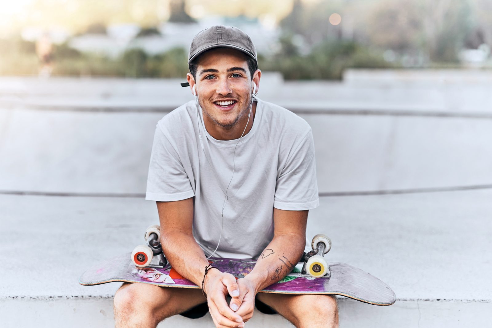 AdobeStock_548186302 Junger Mann sitzt an einem Skatepark, stützt sich auf sein Skateboard ab und lächelt, trägt Kopfhörer und ein graues T-Shirt.