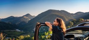 Frau lehnt an Autotür und blickt in gebirgige Landschaft mit Bäumen und Hügeln unter blauem Himmel.