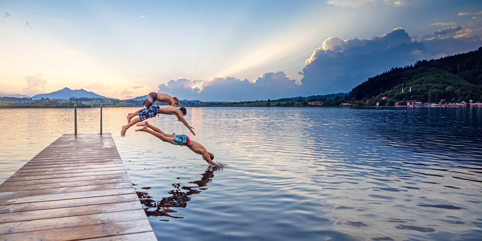 AdobeStock_638446478 Drei Jungen springen vom Holzsteg in einen See, umgeben von Bergen und einem farbenfrohen Sonnenuntergang.